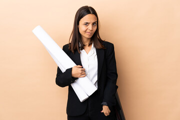 Young architect woman holding blueprints over isolated background having doubts