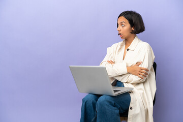 Young mixed race woman sitting on a chair with laptop isolated on purple background doing surprise gesture while looking to the side