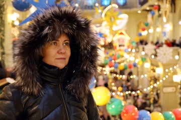 Happy woman in fur hood on background of Christmas decorations in the shopping mall. New Year holidays, defocused view to festive lights and walking people