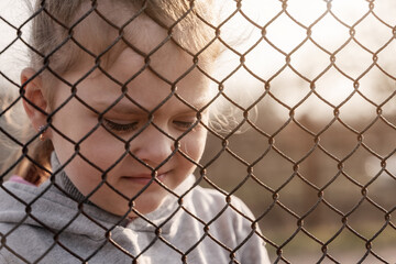 Little girl with a sad look behind a metal fence, social problems, raising children in orphanages
