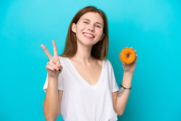 Young English woman holding a donut over isolated blue background smiling and showing victory sign