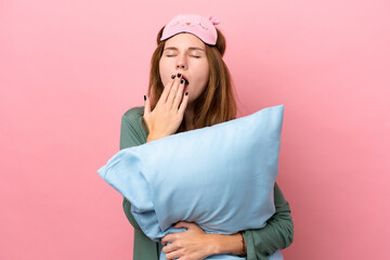 Young English woman in pajamas isolated on pink background in pajamas and holding a pillow and yawning - obrazy, fototapety, plakaty