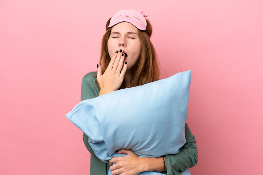 Young English woman in pajamas isolated on pink background in pajamas and holding a pillow and yawning