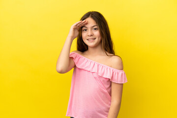 Little caucasian girl isolated on yellow background saluting with hand with happy expression