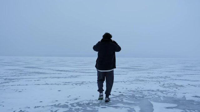 A young lonely guy walks on frozen ice in snowy windy weather. Back view.