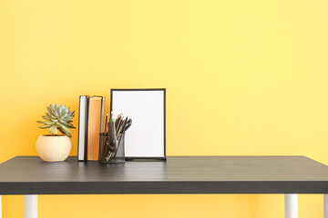 Workplace with stationery holder, houseplant, books and blank photo frame near color wall