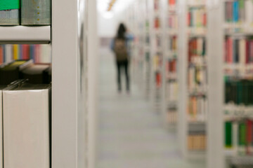 Figura feminina, de costas, andando por corredor de livros de uma biblioteca. Destaque de dois...