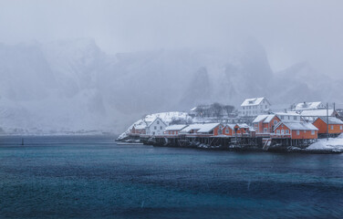 Misty landscape. Winter cloudy day. Traditional Scandinavian-style orange wooden houses stand on panton. Mountains in heavy haze are in the background. Emerald water. Scandinavia.