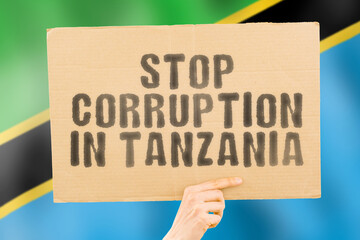 The phrase " Stop Corruption in Tanzania " on a banner in men's hand with blurred Tanzanian flag on the background. Forbidden. Prevent. Wealth. Offence. Corruptness. Economy. Corruptible. Politics