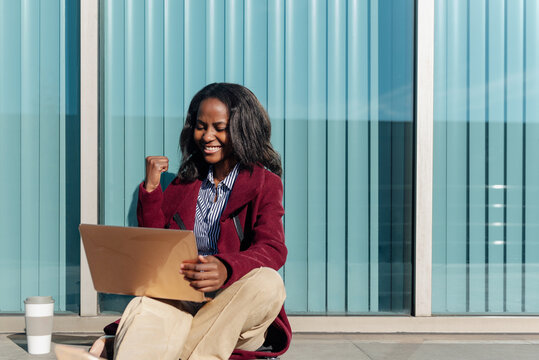 young african female student celebrating the success of her university exams. Black girl using a laptop sitting on the street.