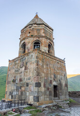 Fototapeta na wymiar Stone walls of Gergeti Trinity Church bell tower, blue morning sky, green caucasiam mountains in the background