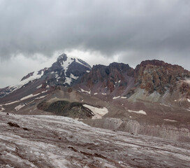 Fototapeta na wymiar Gergeti glacier and mount Kazbek in Georgia, grey thunder sky with clouds, grey and brown stones and rocks, and white stone on the slopes