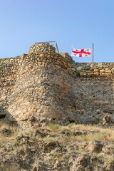 Obraz premium Georgian white and red flag with cross flying in the wind atop the medieval fortress in the town of Gori, Georgia. Ancient yellow, grey and brown stone walls and tower, dry yellow grass, blue sky