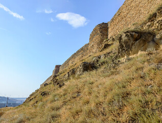 Medieval fortress in the town of Gori, Georgia. Ancient yellow, grey and brown stone walls and towers, dry yellow grass, blue sky with clouds