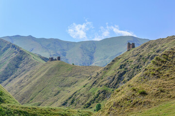 old fortress towers on the mountain road to Shatili, Khevsureti, Georgia. Brown and grey old mossy stone walls, green trees and dry grass on mountain slope, blue sky with clouds.