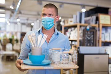 Young man client buyer in face mask choosing variety accessories and tableware in modern shop for...