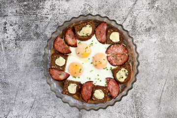 Breakfast - fried eggs with sausage and rye bread in a glass baking dish on a dark grey background. Top view, flat lay
