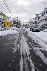 Small one way snow covered street with the center partially plowed and cars parked on both sides in...