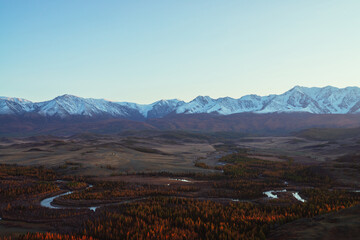 Awesome autumn landscape with mountain river in forest valley and great snow-covered mountain range in red sunset sunshine. Spectacular view from hill to snowy mountains and autumn valley in sunlight.