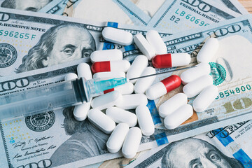 A medical syringe laying next to some pills on top of some hundred dollar bills money cash drugs