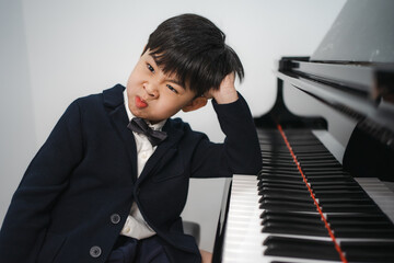 Lazy little boy in black suit sitting at grand piano makes funny face. Cute boring kid pianist...