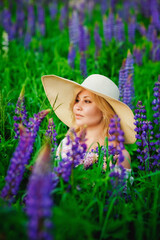 Portrait of a beautiful plump blonde in a large straw hat, a girl with a bouquet of lupines in her hands, posing in a blooming field.