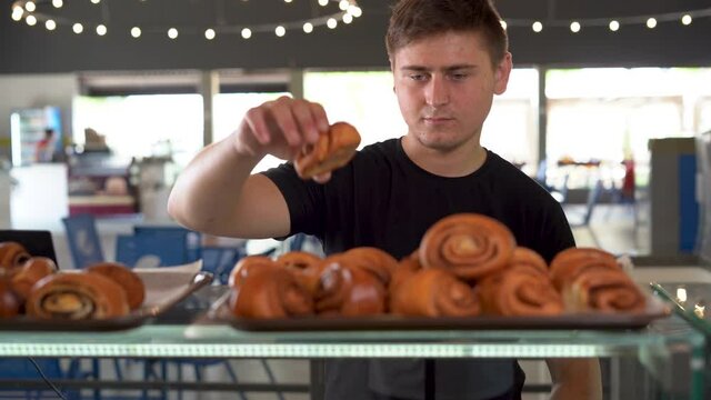 There are cinnamon rolls on the display at the bakery, a Caucasian male shopper picks out a dessert and grabs one roll.