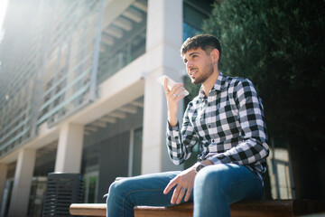 young, handsome guy sitting on a bench and sending an audio note with his phone