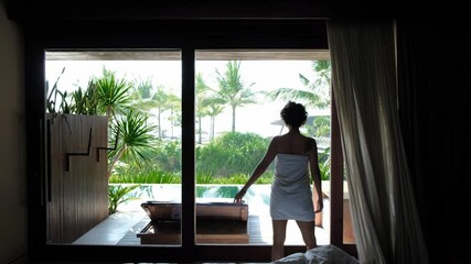 Successful woman covered with towel opening drapes on a tropical luxury villa with swimming pool and palms