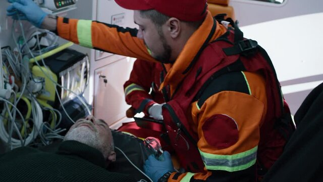 High angle of paramedic putting oxygen mask on senior man near black colleague using tablet while riding ambulance to hospital after emergency