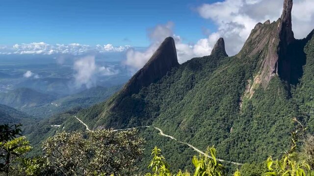 Panning movement showing meandering road beneath the Finger of God peak among Brazilian mountain range called Serra dos Orgaos in Teresopolis with city Rio de Janeiro below in the distance