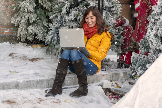 Asian college students wearing yellow jaggers in boots, leaning against pine trees, winter snow clinging to trees and sitting on the floor studying online with a laptop. concept of learning anywhere