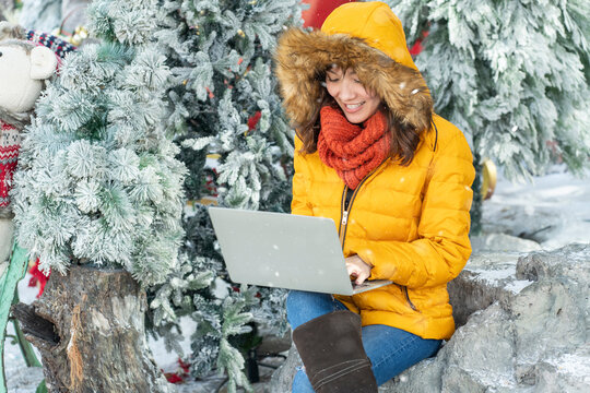 Asian girl wearing a yellow jagger, sitting in a pine garden in winter snow and working online with laptop .Work anywhere concept.