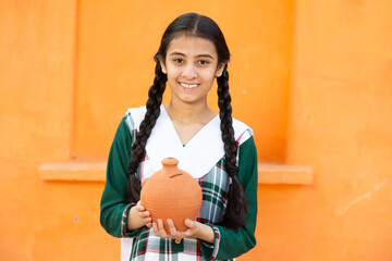 Portrait of happy Indian young girl holidng clay money box or gullak in hand. Smiling braided hair...