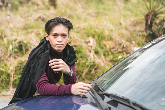 A Young Asian Man In A Purple Sweater And A Big Gold Chain Necklace Leaning On The Hood Of His Blue Car.