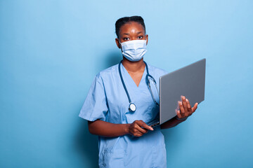 African american physician assistant with protective face mask against coronavirus analyzing medical expertise on computer working in studio with blue background. Health care service. Medicine concept