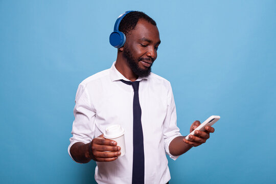 Relaxed man with wireless headphones browsing social media on smartphone while holding cup of coffee to go. Businessman on a break listening to music on over the ear headset looking at smart phone.