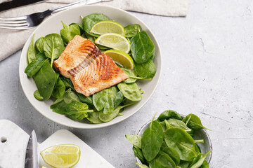 Grey bowl with fresh green raw spinach and fried red salmon, lime slices on grey concrete surface