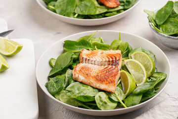 Grey bowl with fresh green raw spinach and fried red salmon, lime slices on grey concrete surface