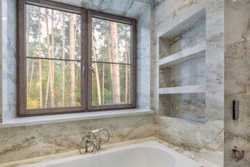 Stylish bathroom with beige marble tiles. The window opens a beautiful view of the pine forest.