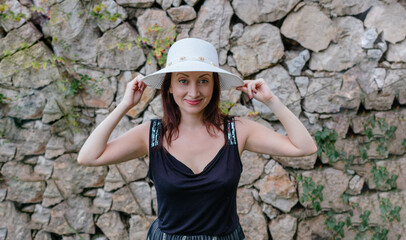 A waist up shot of a charming white woman wearing a hat and smiling with her back to a stone wall.