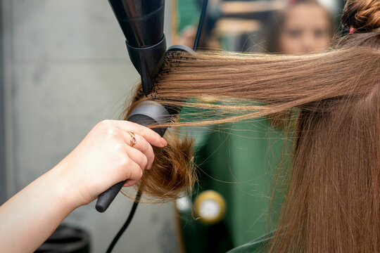 A hairdresser is drying long brown hair with a hairdryer and round brush in a hairdressing salon