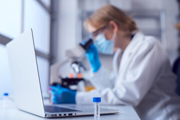 Female Lab Worker Wearing Lab Coat Working On Covid-19 Vaccine With Laptop And Microscope