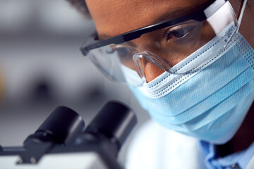 Close Up Of Female Lab Worker Wearing PPE And Safety Glasses Looking Through Microscope