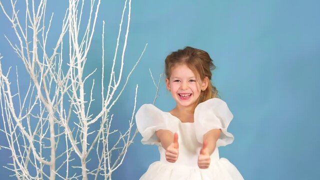 beautiful little girl is dancing and smiling in white fluffy dress on blue background with white tree. Child holiday. Happy childhood. Incendiary dance. Everything is super and cool. Laugh portrait.