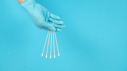 Hand holding swab sticks or cotton sticks. It is wearing blue medical gloves or latex gloves on blue background.