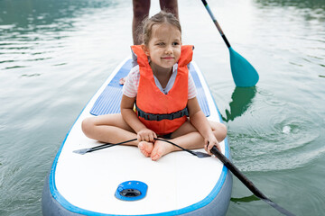 Close up of cute caucasian girl in  swim life vest swimming and her father  on SUP board. Family paddleboarding on the lake on summer day. Active leisure with kids. Family local getaway concept - obrazy, fototapety, plakaty