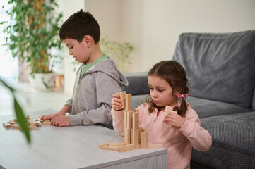 Creative European little girl sitting next to her brother and focused on construction of complex tall structure from wooden blocks, . Fine motor skills, concentration and educational leisure concept