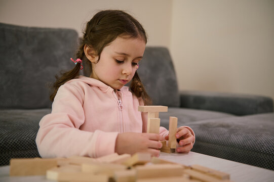 Beautiful creative Caucasian child, little girl playing board game, concentrated on building constructions with wooden blocks bricks. Hand movement control and building computational skills concept.