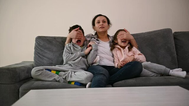 Middle-aged woman, loving mother sits on the sofa and hugs her children, watches TV together when she suddenly covers their eyes with her hands